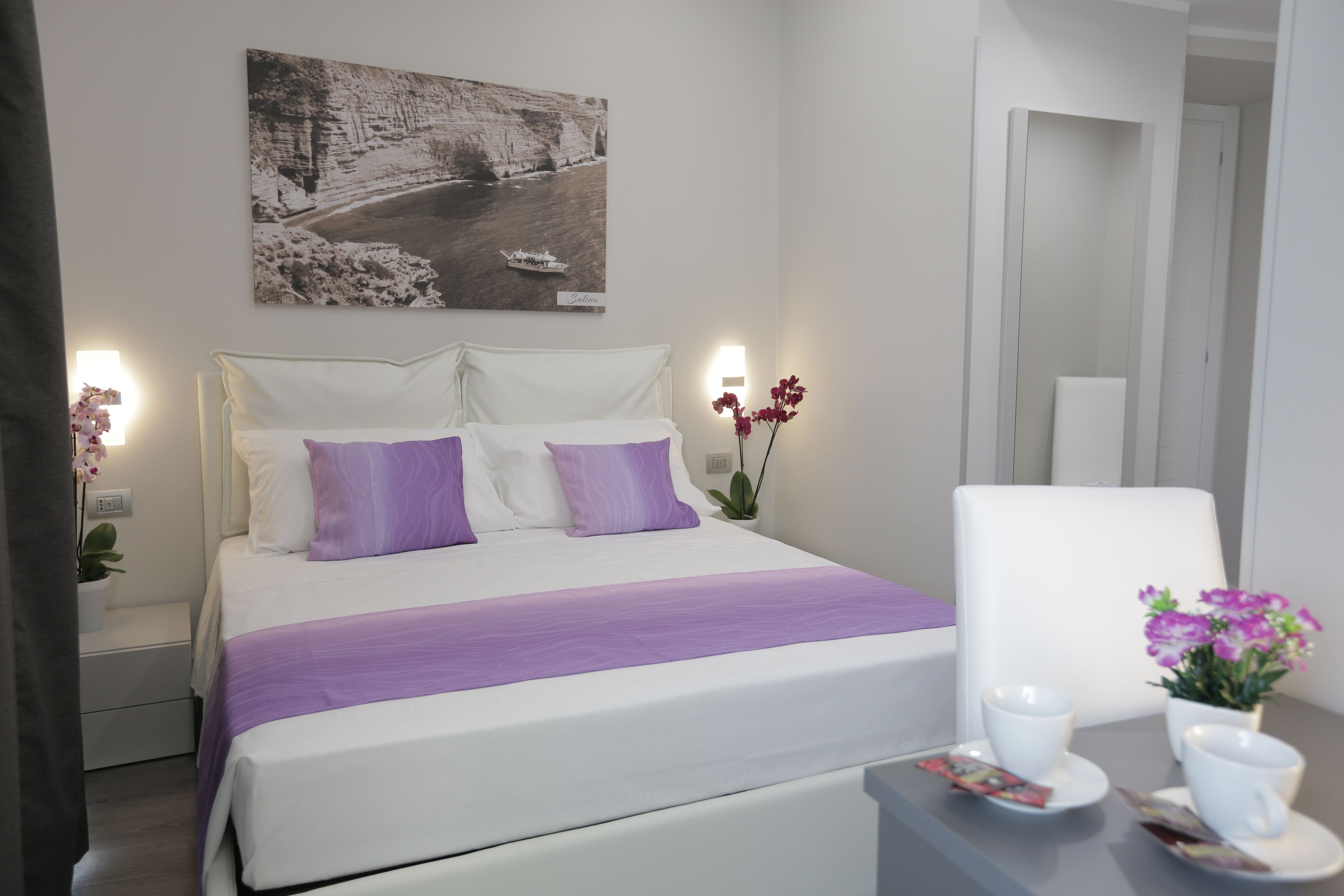 A neatly arranged hotel room with a bed, a chair, a table with a cup and saucer, and a wall-mounted picture.