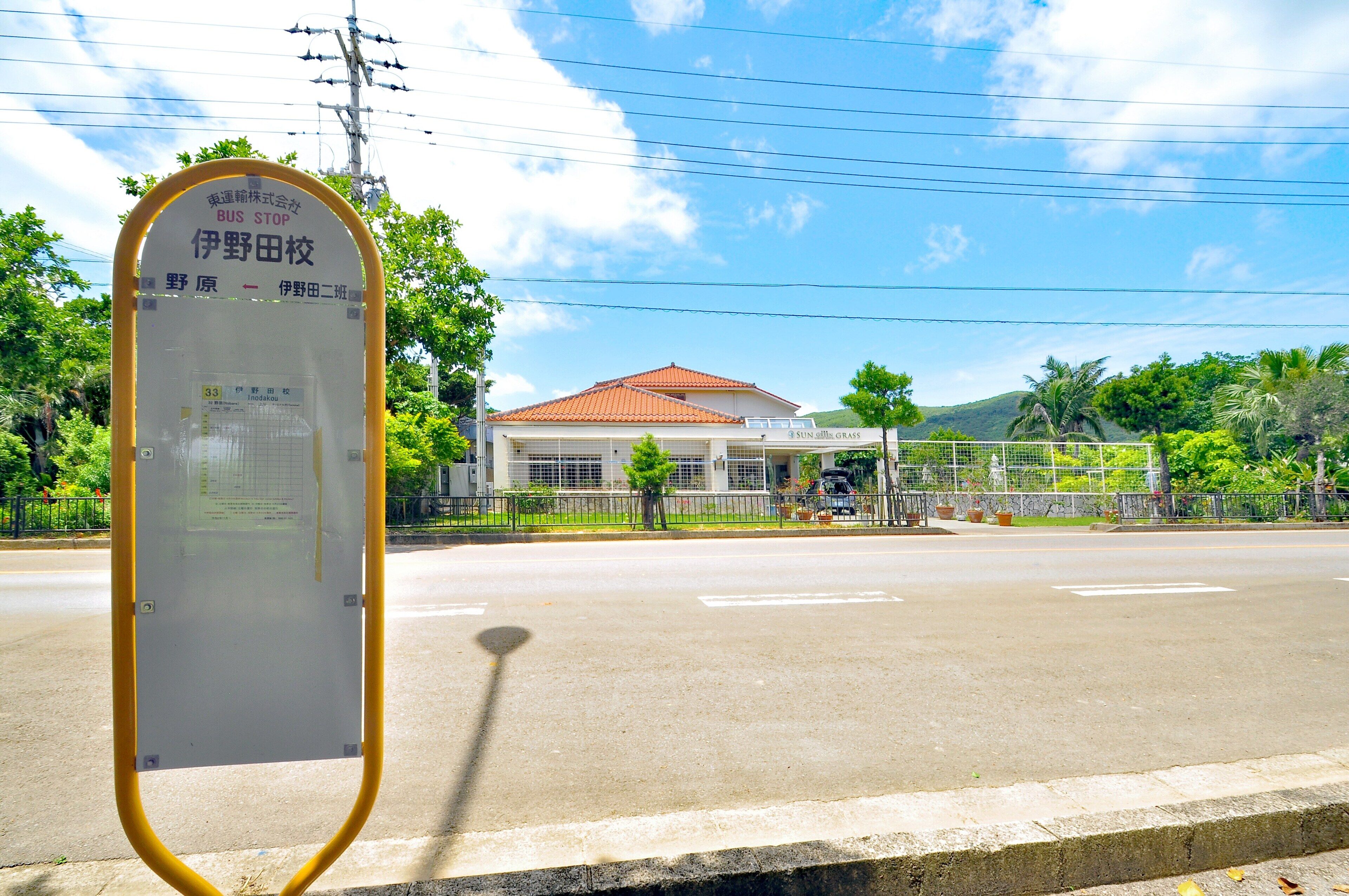 A bus stop sign with route information, a building with a red roof, and a clear sky.