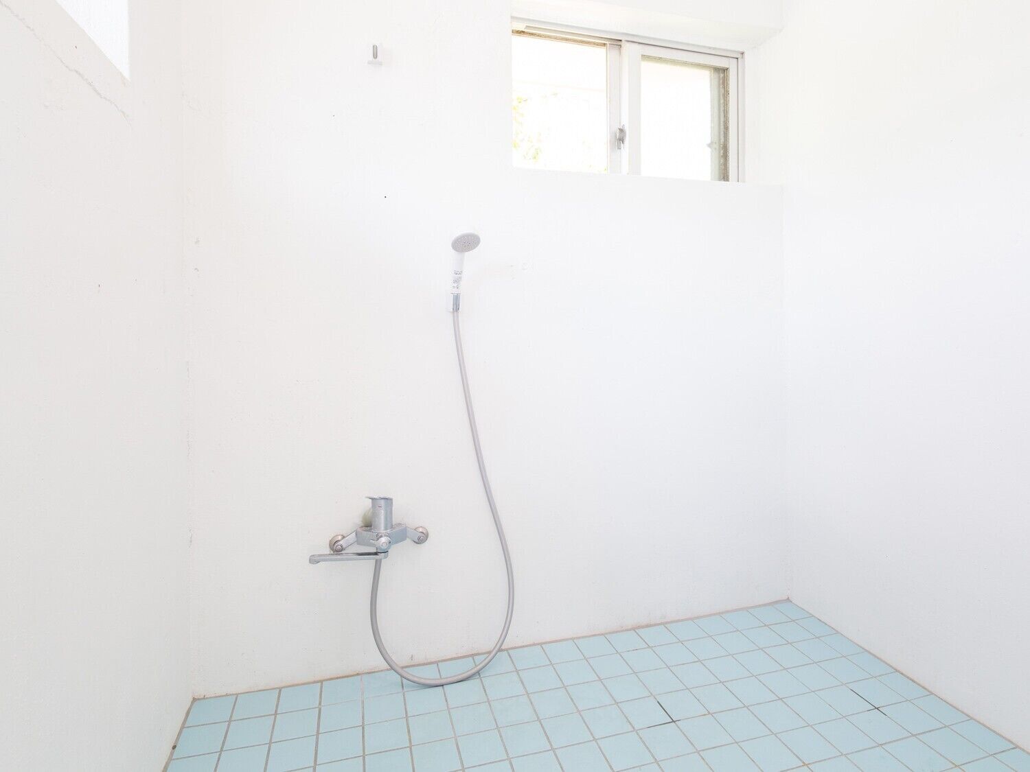 A small, white bathroom with a shower, a window, and light blue tiled flooring.