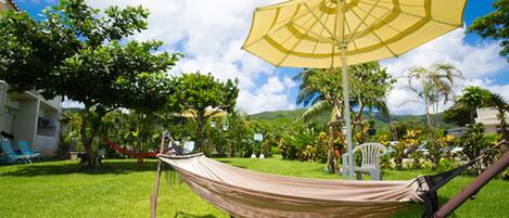 A hammock under a yellow umbrella in a grassy area with trees and a building in the background.