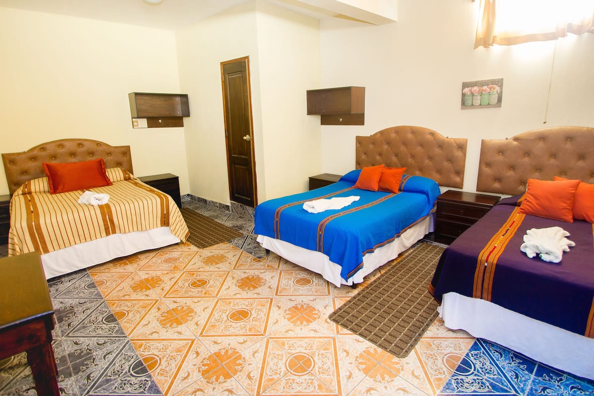 A hotel room with three beds, each with different colored bedding. There are two bedside tables, a door, and a wall-mounted TV.