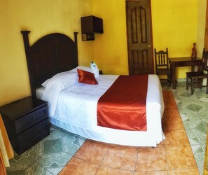 A bed with white and red bedding, a nightstand, a wooden door, a chair, and a small table. - Hotel Santa Maria (Panajachel)
