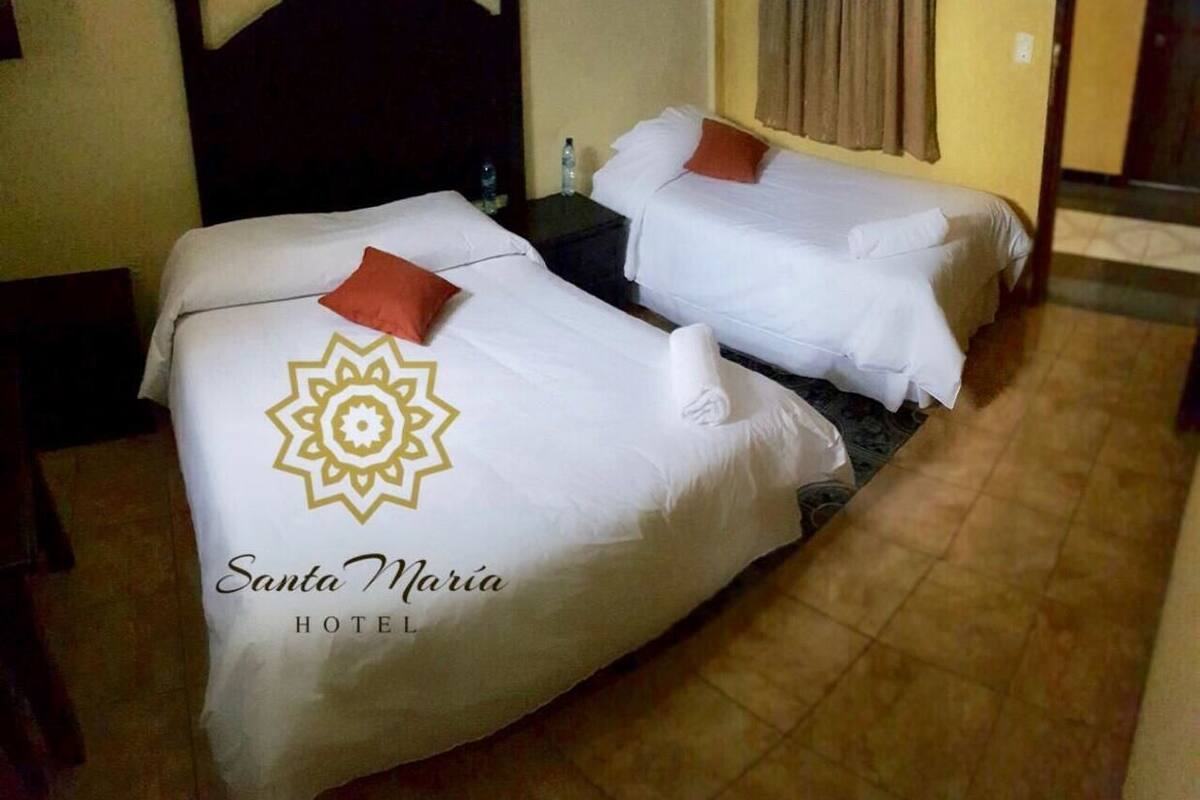 Two single beds with white bedding and orange pillows, a wooden headboard, a small table with a water bottle, and a tiled floor.