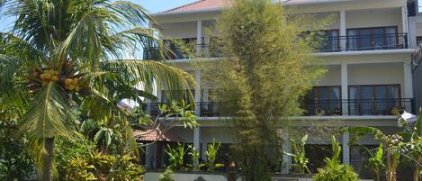 Un edificio de dos pisos con balcones, rodeado de vegetación tropical, incluyendo palmeras y plantas de plátano.