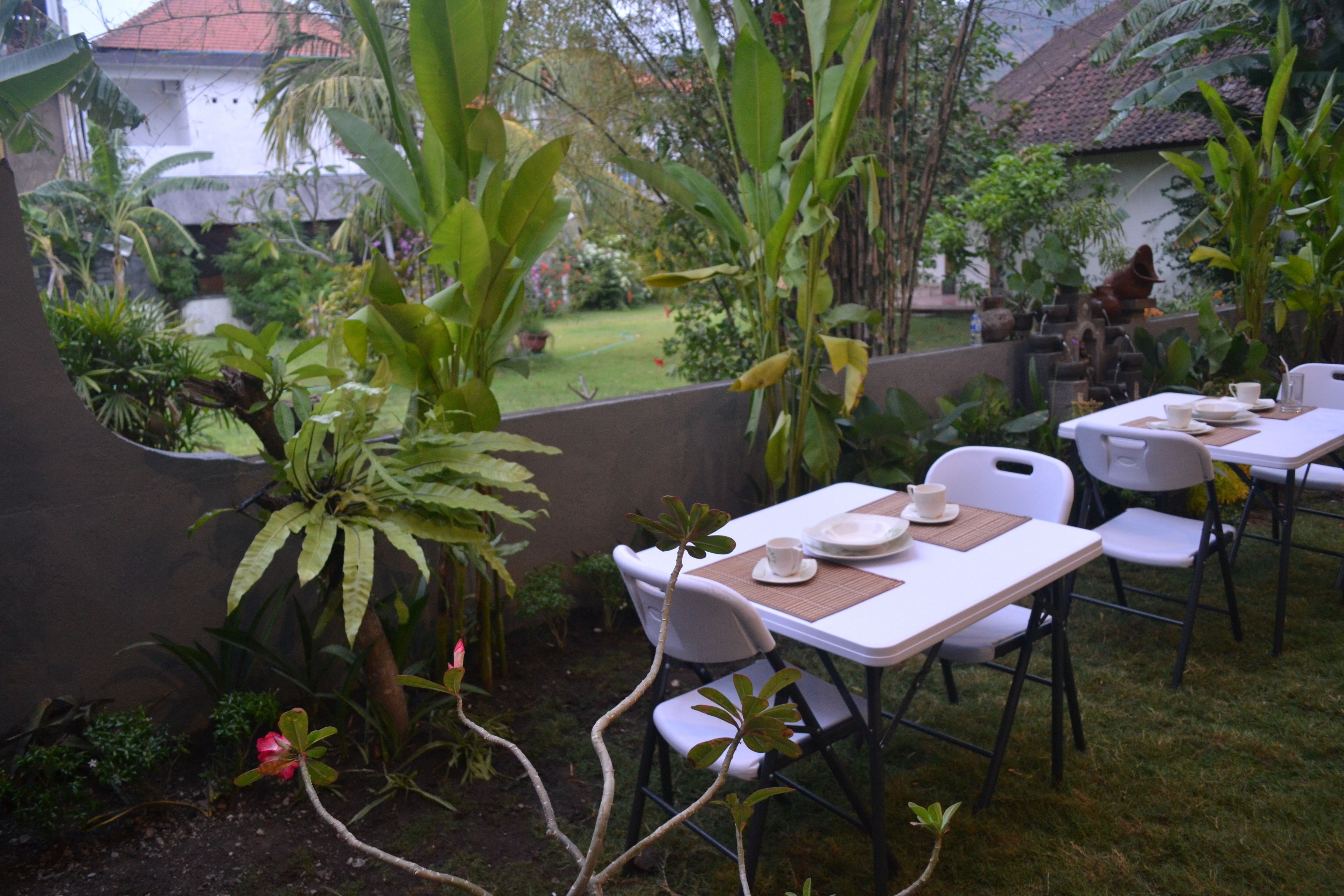 Un patio al aire libre con mesas y sillas, rodeado de abundante vegetación y vista a un edificio residencial al fondo.
