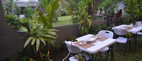 Un patio al aire libre con mesas y sillas, rodeado de abundante vegetación y vista a un edificio residencial al fondo.