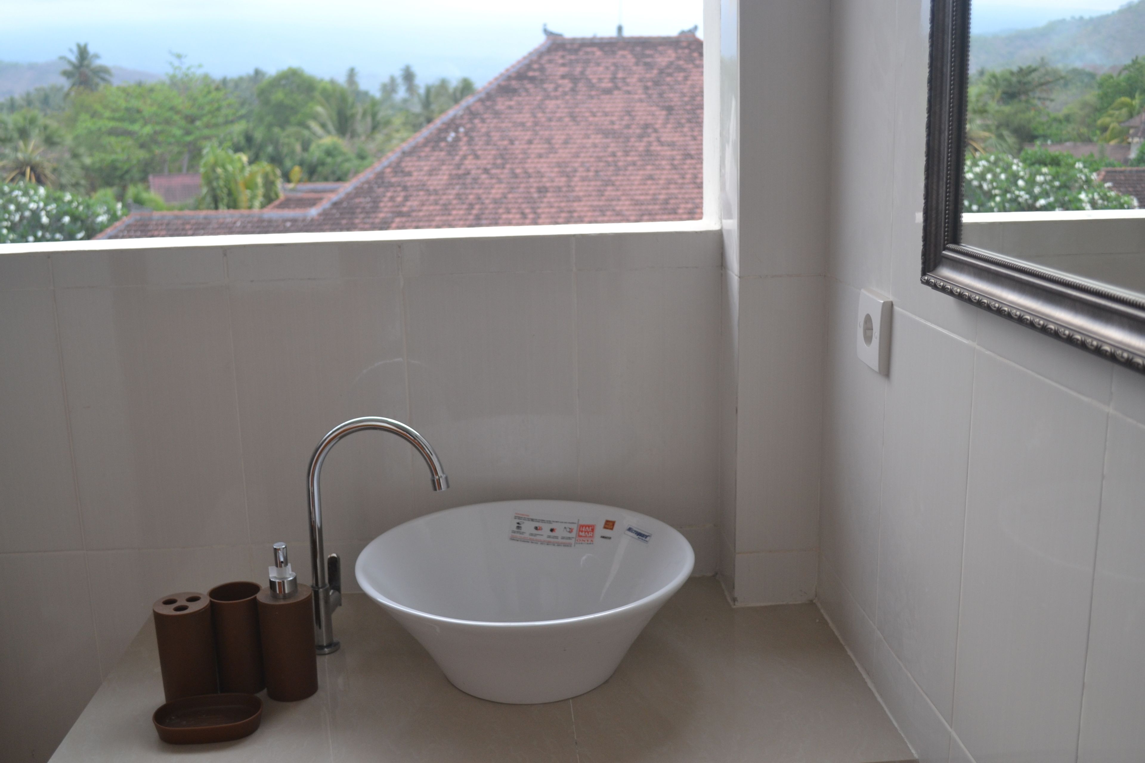 A bathroom with a white sink, a mirror, and a view of a red-roofed building and greenery outside.