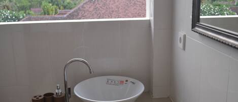 A bathroom with a white sink, a mirror, and a view of a red-roofed building and greenery outside.