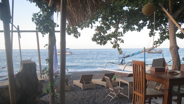 Un área de descanso frente a la playa con sillones y una mesa, con vista al mar y barcos al fondo.