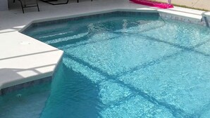 Ein Schwimmbecken mit klarem, blauem Wasser, umgeben von einer weißen Terrasse und einer überdachten Veranda mit Sitzgelegenheiten.