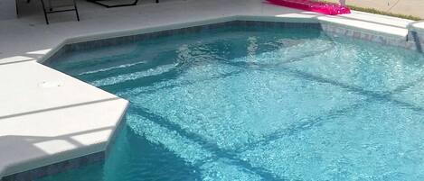 A swimming pool with a clear blue water, surrounded by a white deck and a covered patio area with chairs.