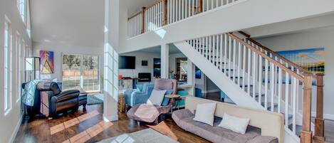 A spacious living room with a high ceiling, a wooden staircase, and a large window allowing natural light.