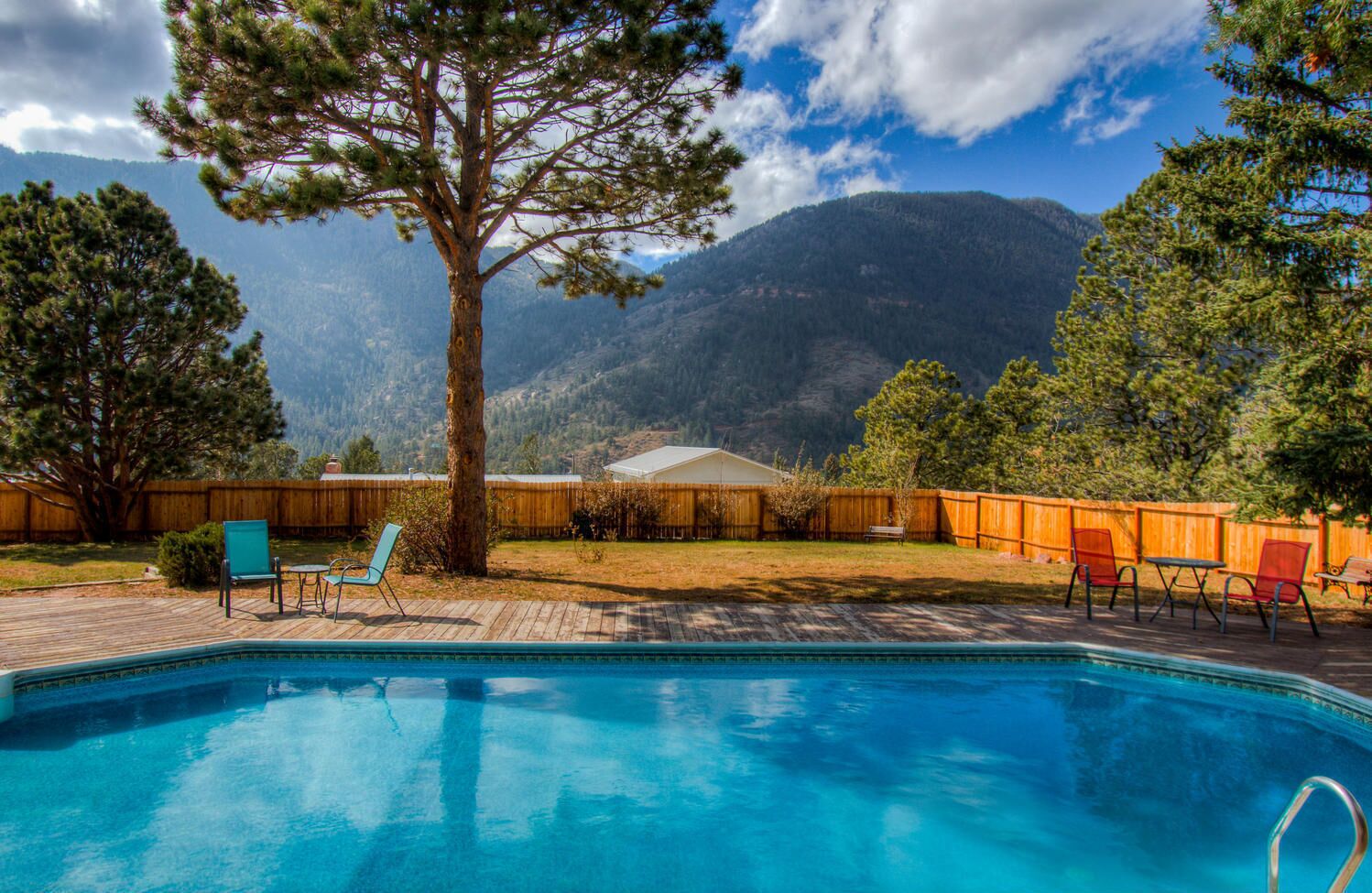 Une piscine dotée d’une terrasse en bois, entourée d’une clôture en bois et d’arbres, avec une vue sur les montagnes en arrière-plan.
