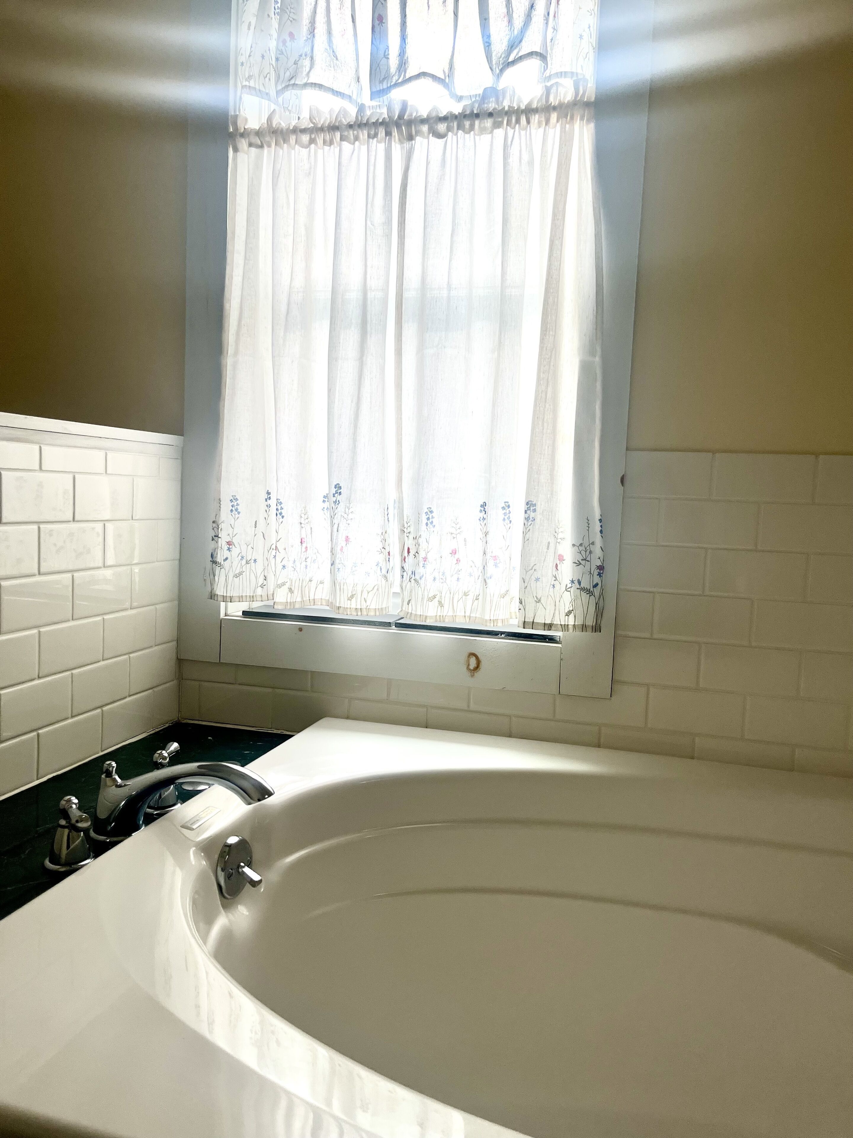 A bathroom with a white bathtub, a window with floral curtains, and white tiled walls.