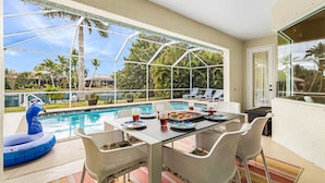 Un espace repas aménagé au bord de la piscine, avec une table pour deux, entouré de chaises en osier blanc, donnant sur une piscine avec des chaises longues et des parasols.