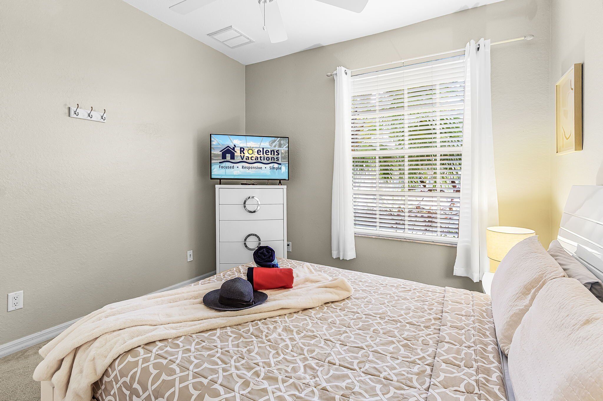 A bedroom with a bed, a TV on a stand, a window with blinds, and a wall-mounted light.