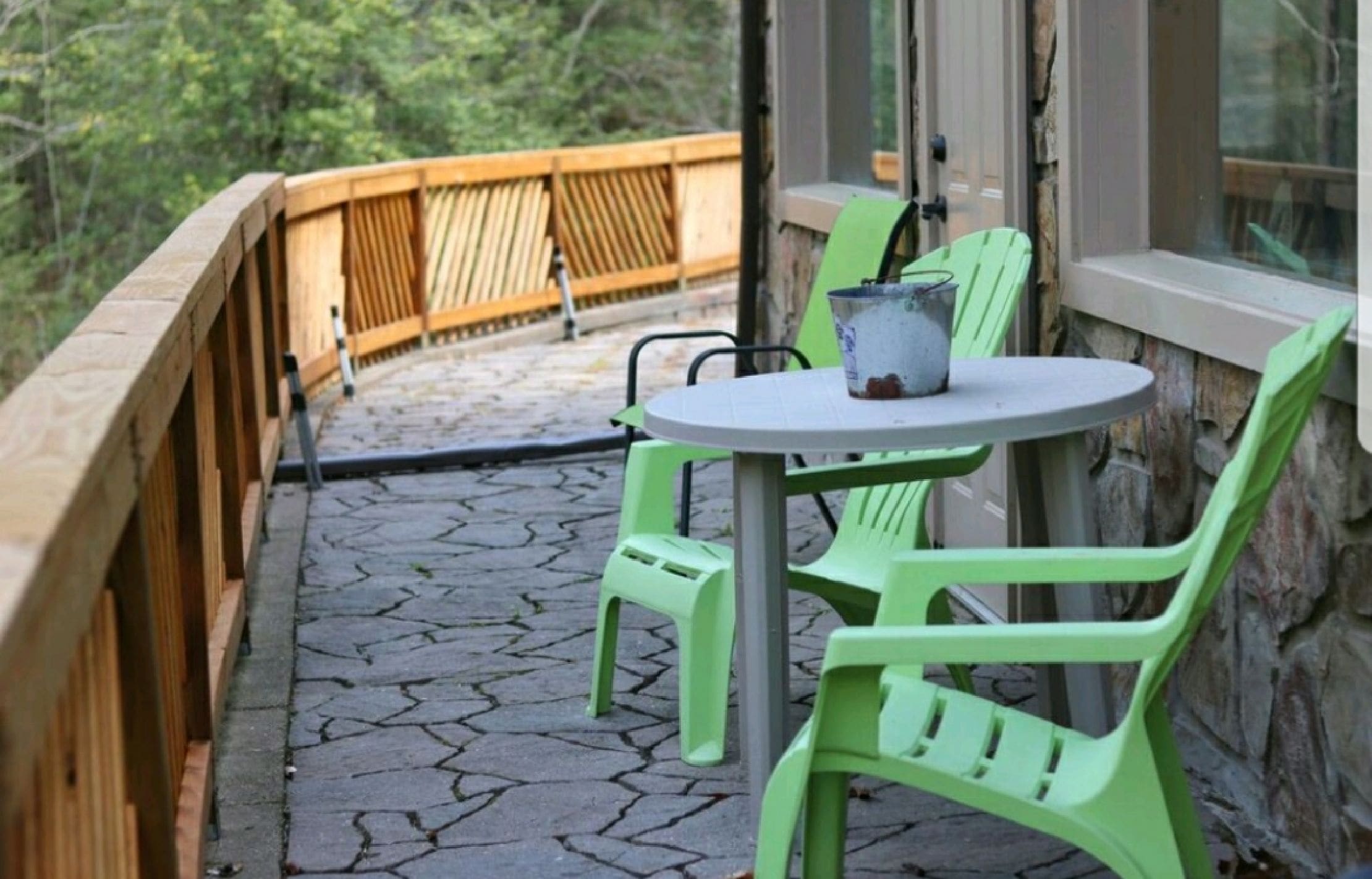 Un patio con una mesa redonda y dos sillas verdes, una cerca de madera y un muro de piedra.