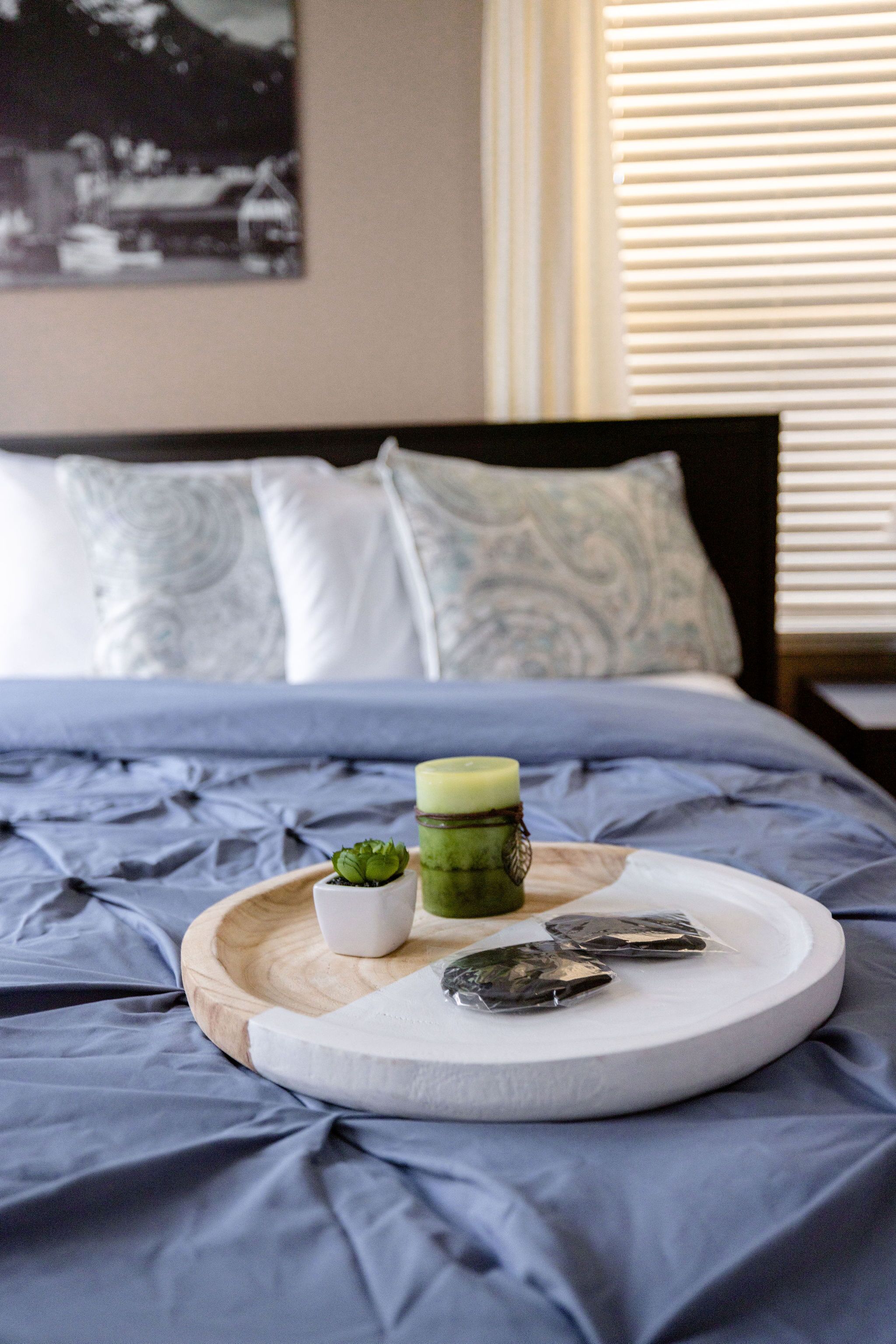 A neatly made bed with a blue comforter, a wooden tray with a candle, a small plant, and two wrapped items on a white pillow.