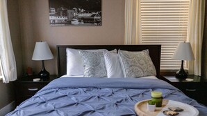 A neatly made bed with a blue quilt, two bedside lamps, a tray with food and a candle, and a black and white photo on the wall. - Comfort and Convenience in Downtown Sitka - Suite A (Sitka)
