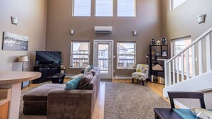 A spacious living room with a large brown sofa, a television, and a staircase leading to an upper level. - Comfort and Convenience in Downtown Sitka - Suite A (Sitka)