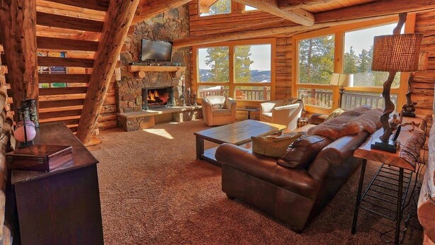 L’intérieur d’une cabane en rondins, avec une cheminée en pierre, des meubles en bois et de grandes fenêtres donnant sur le paysage extérieur.