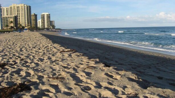 A sandy beach with ocean waves, tall buildings in the background, and a clear sky.