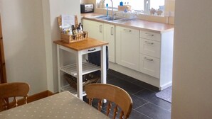 A kitchen with white cabinets, a wooden table, and a window with blinds. - Modernised traditional cottage, Sleeps 5,short walk to beaches, town, amenities  (Tenby)
