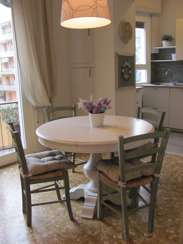A dining area with a round table, four chairs, a vase with flowers, a hanging lamp, and a view of a balcony with plants.