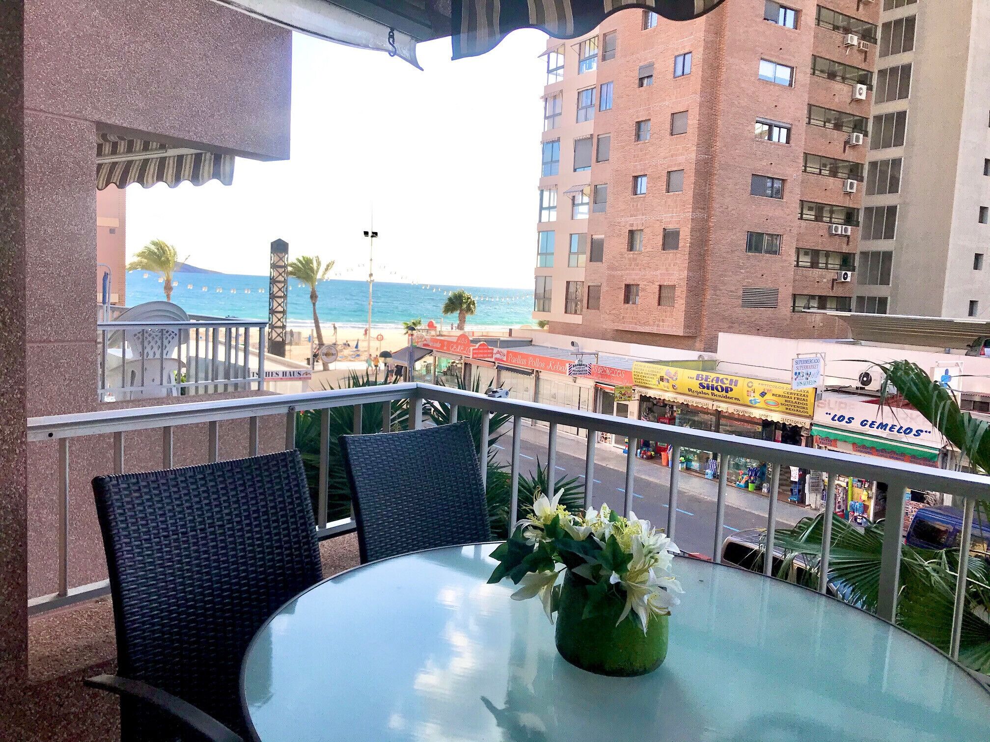 Een balkon met een glazen tafel en stoelen, met uitzicht op een strand en een winkelgebied.