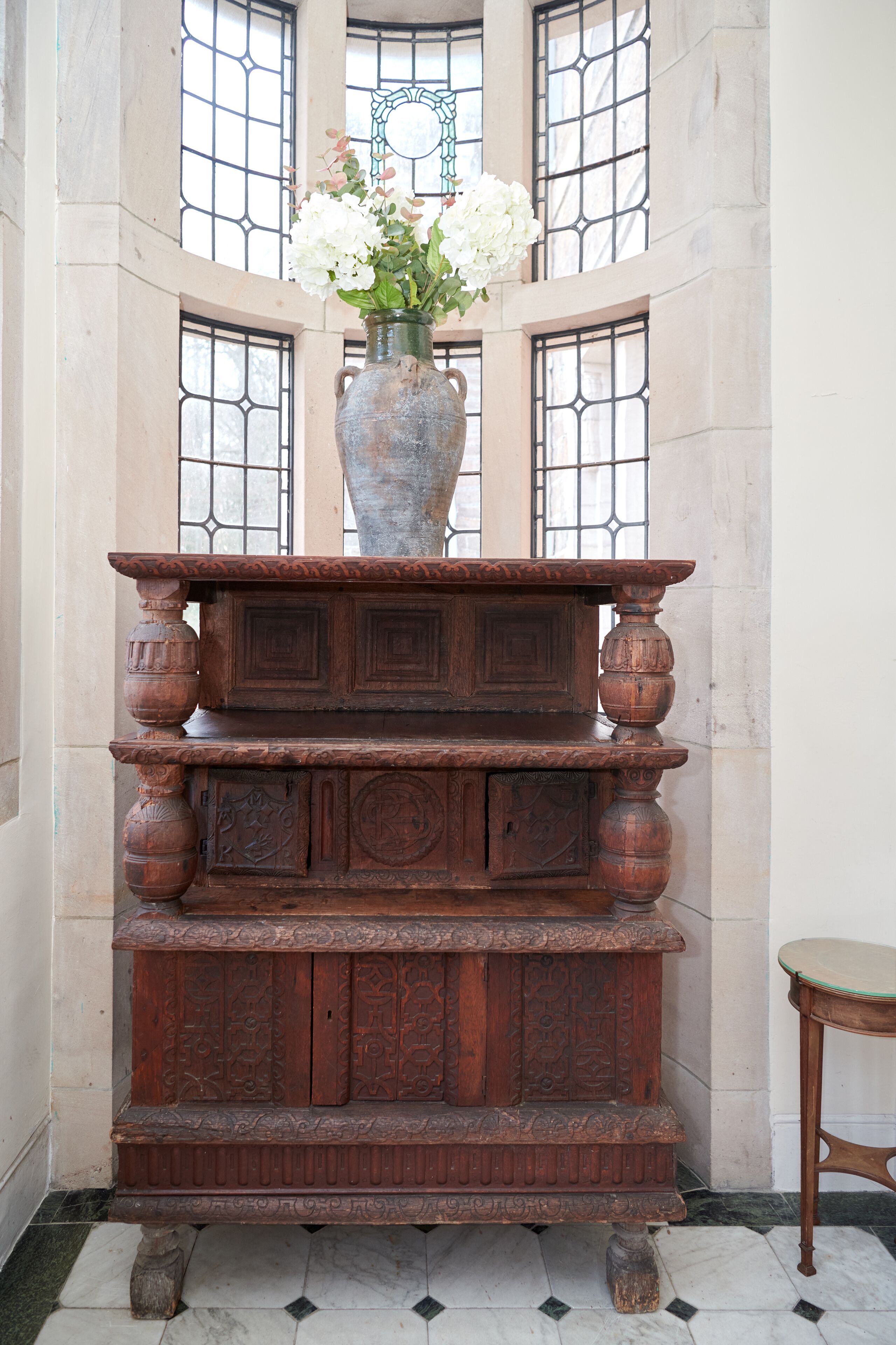 Ein Holzkabinett mit aufwendigen Schnitzereien, eine Vase mit weißen Blumen und ein Fenster mit dekorativem Gitter.