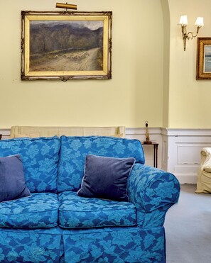 Una habitación con un sofá azul estampado, un cuadro de un paisaje enmarcado y una lámpara fijada a la pared.
