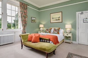 Een slaapkamer met een bed, een groene fauteuil, een radiator, een raam met gordijnen en een deur.