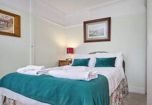 A neatly made bed with a teal bedspread and white pillows, a wooden nightstand with a red lamp, and two framed landscape paintings on the walls. - Classically beautiful large family friendly retreat, situated on the Moray Firth (Moray)