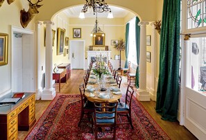 Een eetzaal met een lange tafel gedekt voor een maaltijd, omringd door stoelen, een kroonluchter en een tapijt met een patroon.