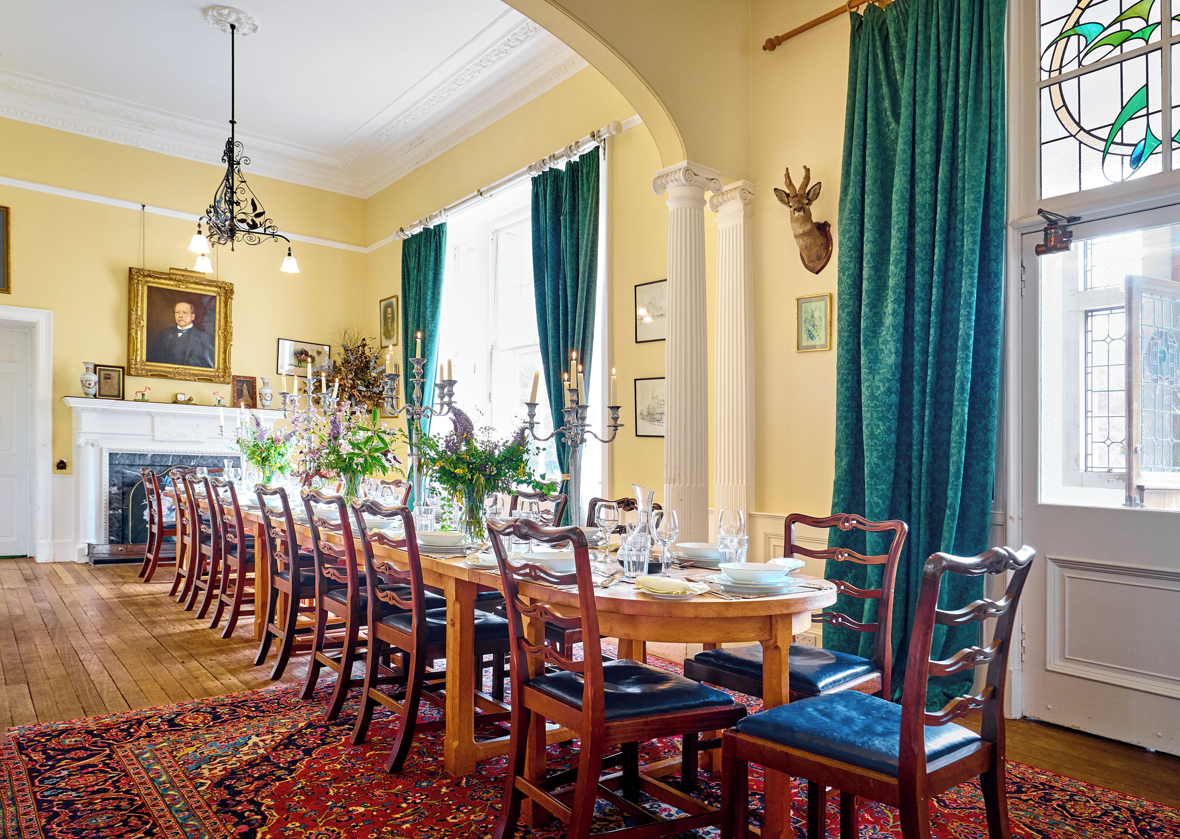 Une salle à manger avec une longue table dressée pour un repas, entourée de chaises, et agrémentée d’une cheminée et d’un lustre.