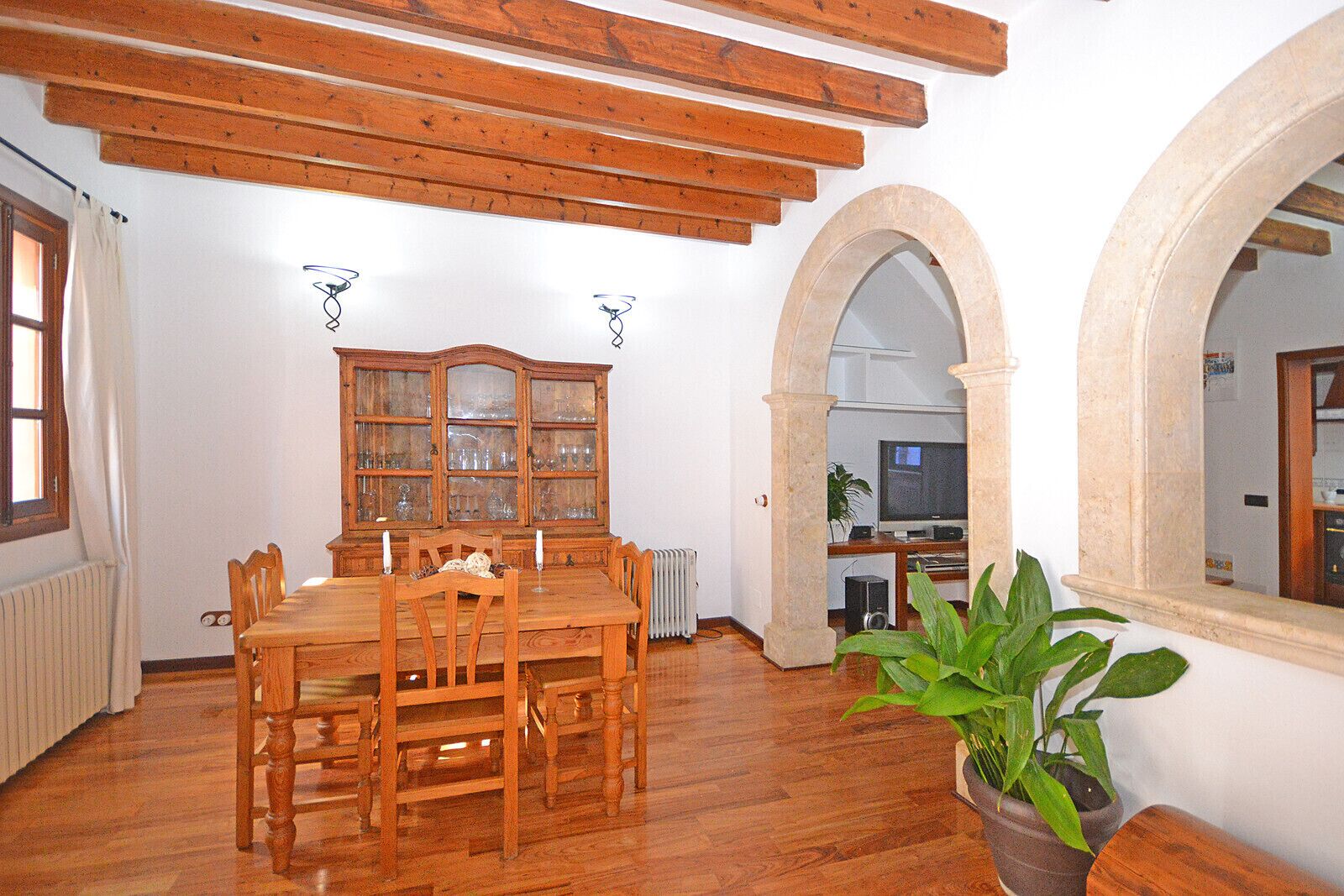 Un comedor con mesa y sillas de madera, un mueble con puertas de vidrio y un radiador. Hay una planta en la esquina y un televisor al fondo.
