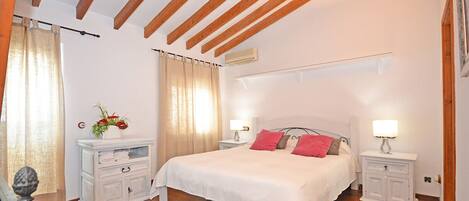 A bedroom with a large bed, wooden beams on the ceiling, white nightstands, and a small bedside table with a lamp.
