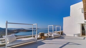 A white outdoor seating area with a view of the sea and distant hills.