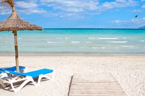 A beach with clear turquoise water, a wooden walkway, and blue lounge chairs under a thatched umbrella.