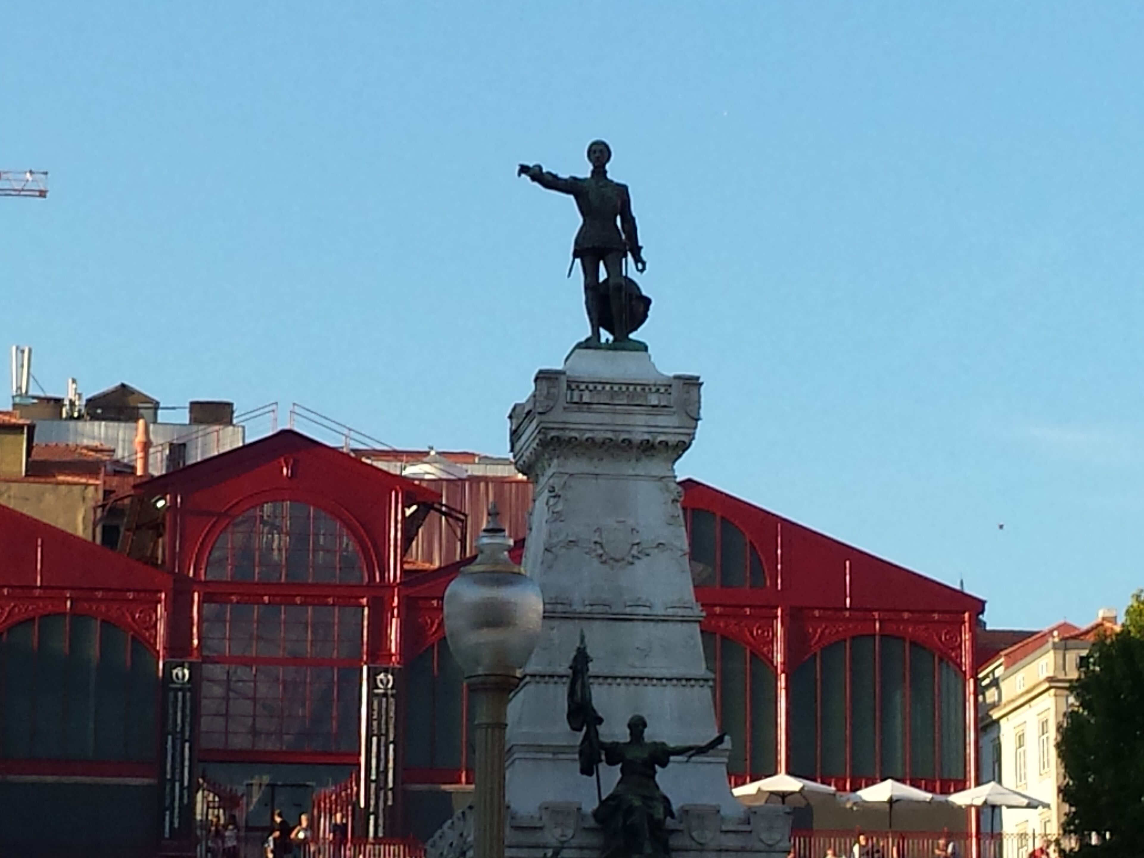 A statue on a pedestal with a figure holding a flag, set against a backdrop of buildings with large windows and red awnings.
