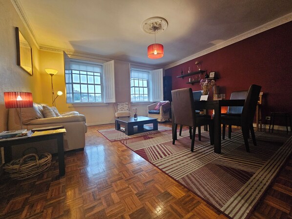 A living room with a sofa, a coffee table, a dining table with chairs, a fireplace, and large windows.