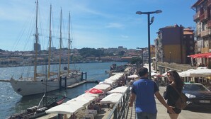 A harbor with a tall sailing ship docked, a bustling waterfront promenade with outdoor seating, and buildings with balconies overlooking the water.