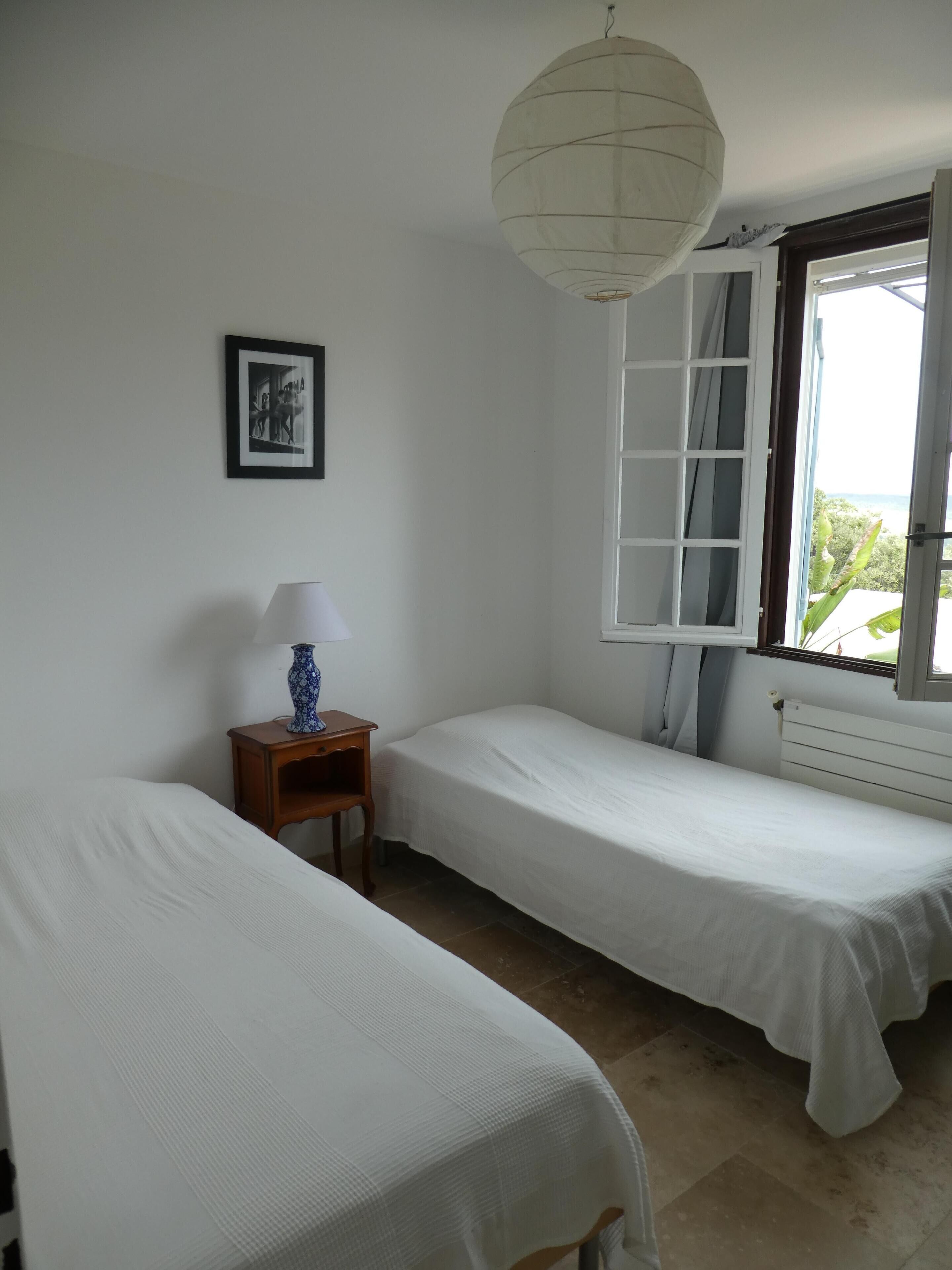 Una habitación con dos camas individuales, una mesita con una lámpara, un cuadro en la pared y una ventana con cortinas.