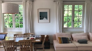 A cozy living room with a dining table, wooden chairs, a sofa with cushions, a framed picture on the wall, and large windows with curtains. - Stylish villa with private heated swimming pool, breathtaking view, free wifi (Lorgues)