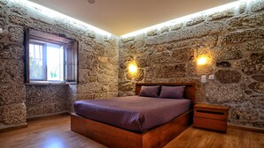 Une chambre avec un mur en pierre, un lit en bois, une table de chevet et une fenêtre laissant entrer la lumière du jour.