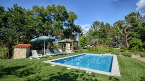 Une piscine entourée d’arbres et d’une structure en pierre, avec des chaises longues et des parasols à proximité.
