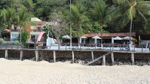 Una proprietà fronte spiaggia con una passerella in legno che conduce a un ristorante con posti a sedere all'aperto, circondato da palme e un muro in pietra.