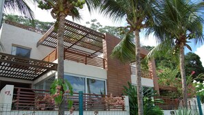 Una moderna casa a due piani con un balcone, immersa in una vegetazione tropicale e palme.