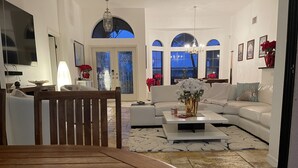 Un saló modern amb una taula de dinar de fusta, sofàs blancs, una taula de centre amb un ram i una finestra gran amb arcades.
