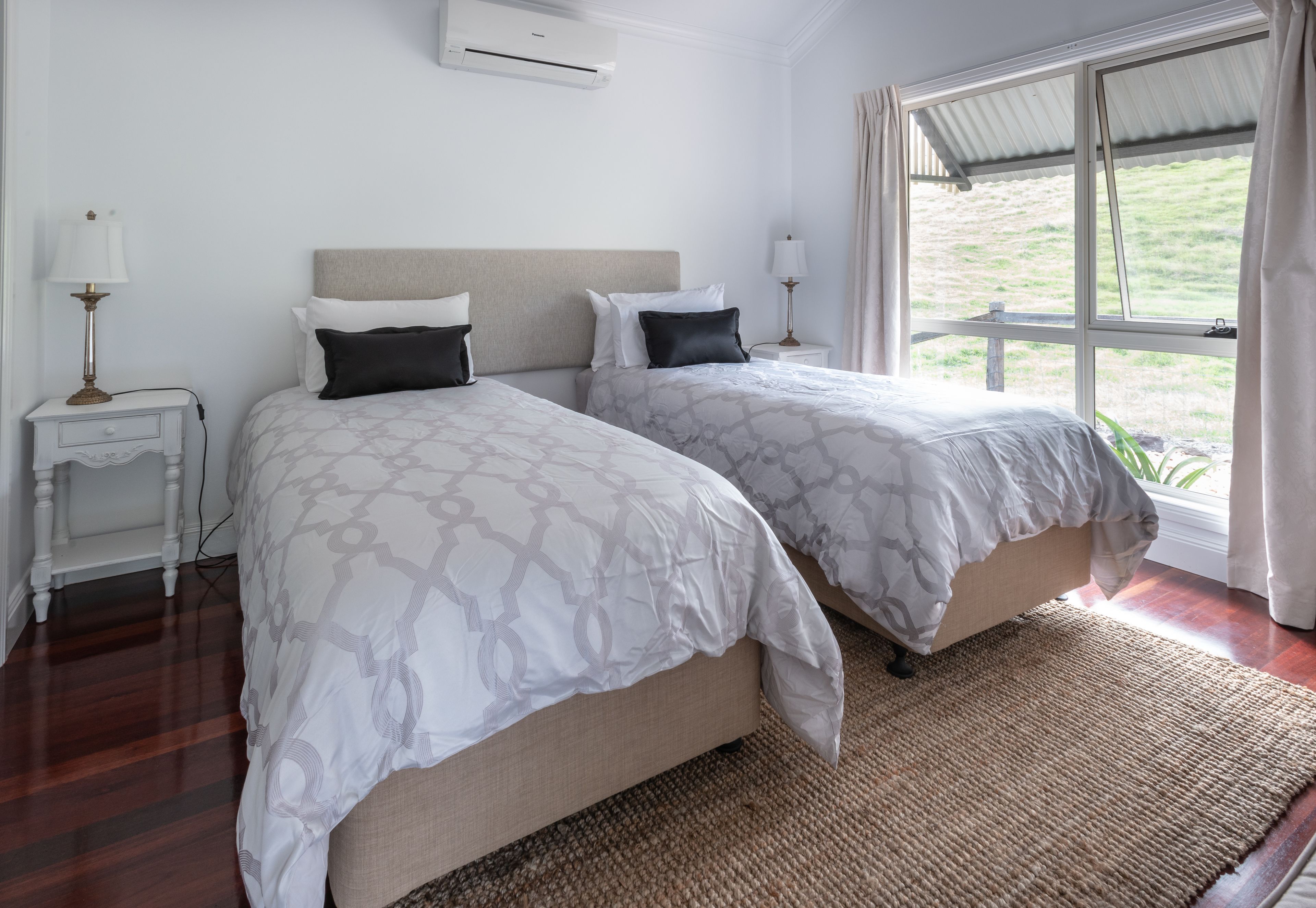 A bedroom with two beds, a window with curtains, a bedside table with a lamp, and a wall-mounted air conditioner.